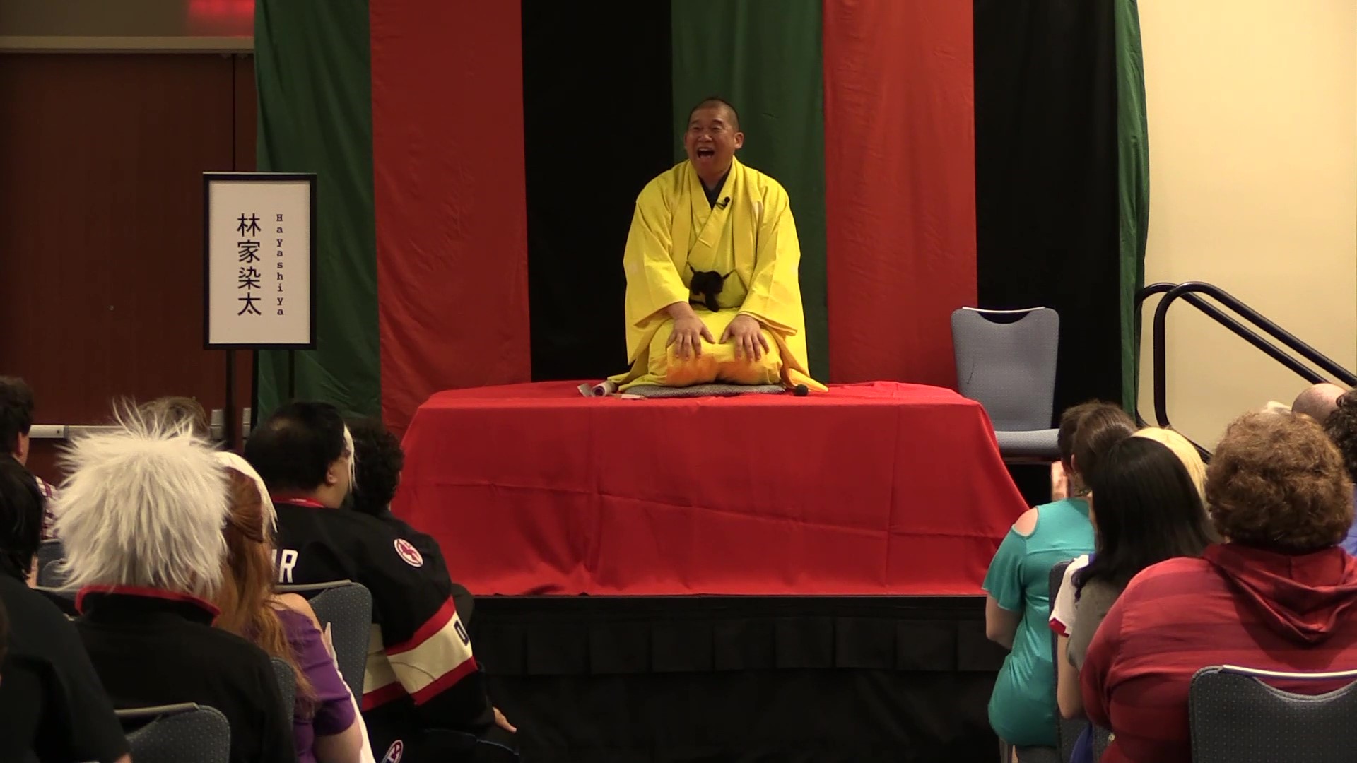 a man kneels on a stage wearing bright yellow traditional Japanese clothing. He is smiling out at an audience.