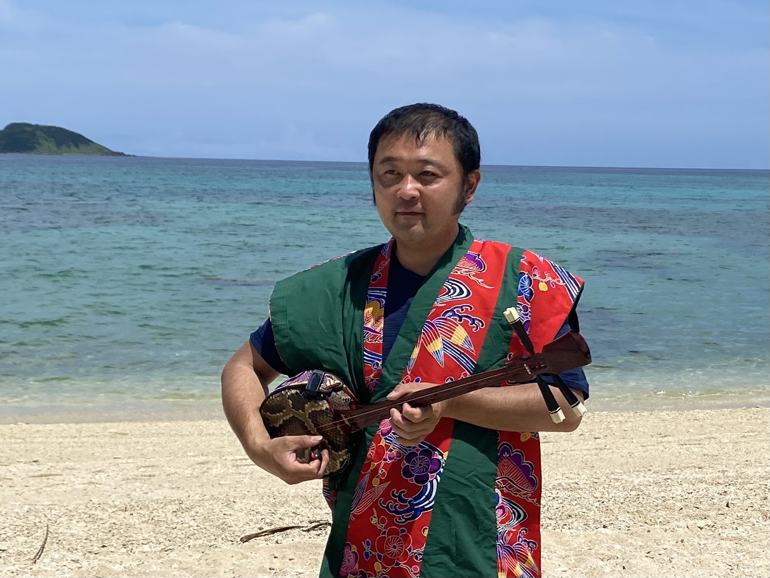 a man in traditional Japanese clothing stands on a sandy beach. He is playing a Japanese instrument called a sanshin.